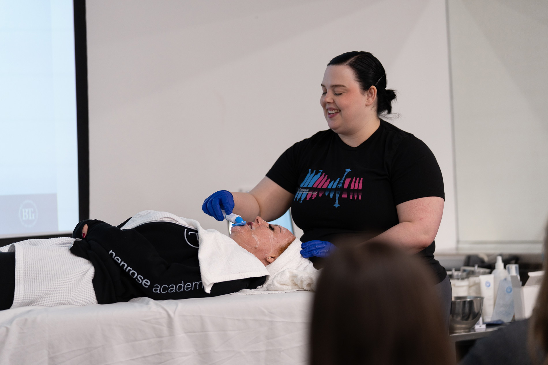 woman in the front of a classroom performing the steps of a facial on female